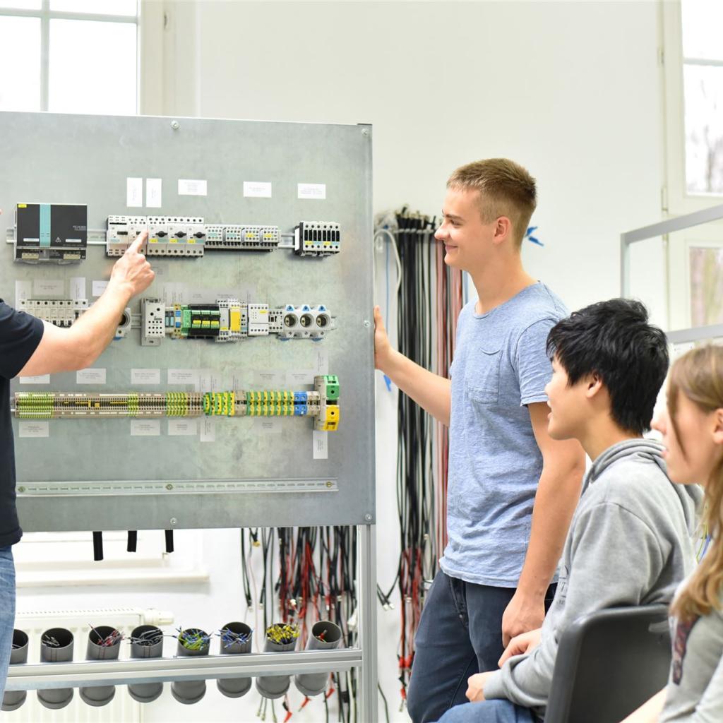 3 Auszubildende und der Ausbilder stehen vor einer Schalttafel 