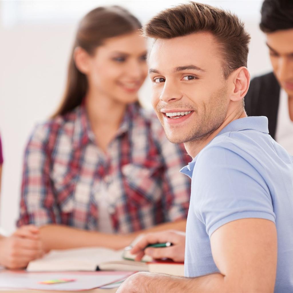 4 Studenten sitzen am Schreibtisch, einer davon dreht sich um und lächelt in die Kamera