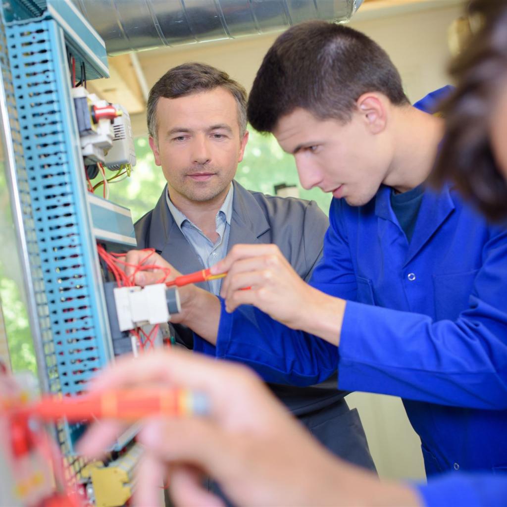 2 junge männliche Elektoniker arbeiten an einer Schalttafel, im Hintergrund der Ausbildungsleiter