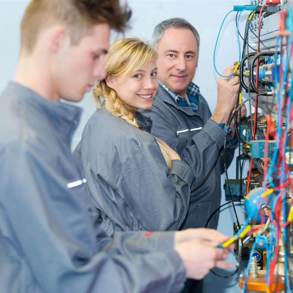 2 Auszubildende, eine junge Frau und ein junger Mann arbeiten an einer Schalttafel, im Hintergrund gibt der Ausbilder Anweisungen